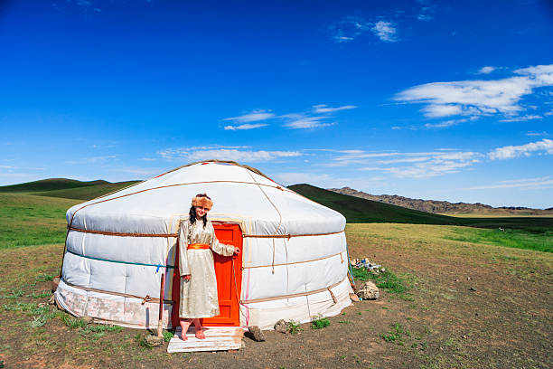 A bright white yurt with an orange door sits on an open grassy plain under a wide blue sky. A woman in a long cream robe with an orange sash and a fur hat stands barefoot on a small wooden step, holding the door and smiling. Gentle rolling hills and rocky outcrops stretch into the distance, creating a peaceful pastoral scene.