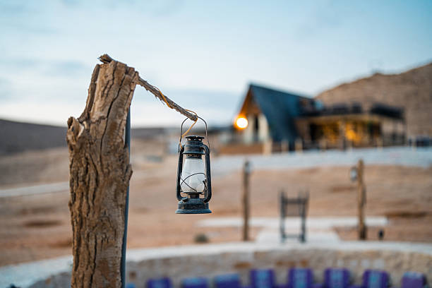 Rustic lantern hanging from a wooden post at Ghamra Camp in Saudi Arabia, with a blurred background of a cabin. Evening light creates a tranquil atmosphere.