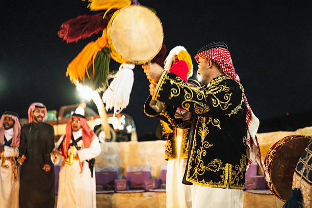 Saudi men performing the traditional Ardah dance at Ghamra Camp's evening entertainment. Performers wearing traditional clothing and headdresses, playing drums. The scene cultural celebration and heritage with a warm glow during the event at night.