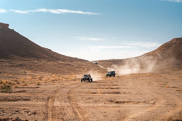 Tourist buggies navigating a dirt path in the expansive Saudi Arabian desert under a clear blue sky. Dust trails follow the vehicles, highlighting the arid landscape and adventurous atmosphere of the desert setting.