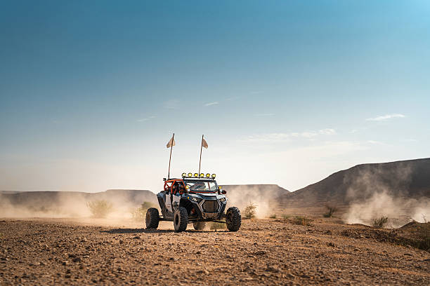 Arab tourists riding in an off-road buggy, driving along a dirt path in the sandy Saudi Arabian desert. The buggy kicks up dust as it moves across the arid terrain under a clear sky during the day. Adventure and exploration concept.