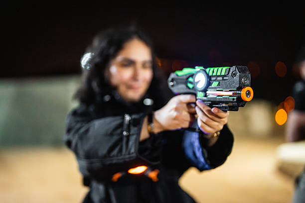 Young Middle Eastern female focusing laser tag gun at Ghamra Camp. Smiling, holding the laser tag equipment with illuminated features. Enjoying recreational activity in a nighttime setting. Dark background creates high contrast on the subject, capturing a fun moment.