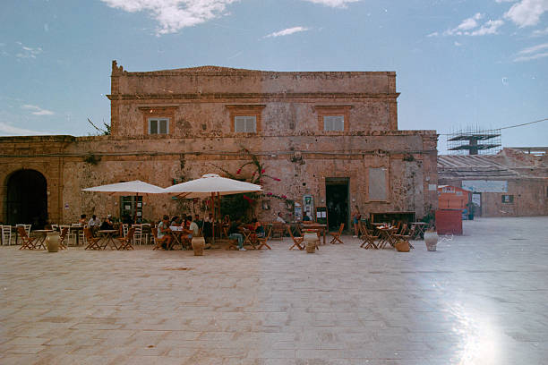 Marzamemi, Sicily, August 29th, 2021: People enjoying outdoor dining at a cafe in the quaint main piazza of the historic fishing village Marzamemi, Sicily, Italy. Scanned from analog film