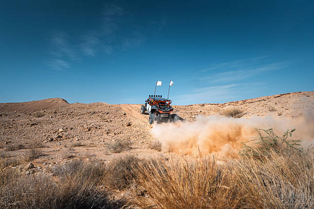 An Arab tourist driving a buggy, creating a dust cloud, on a dirt path through the Saudi Arabian desert. The sun shining, casting shadows across the arid landscape. Clear blue sky above, enhancing the sense of adventure and vastness.