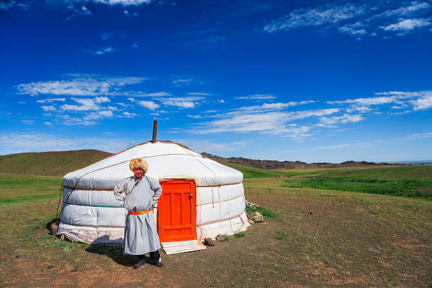 A person wearing traditional clothing and a fur hat stands with hands on hips outside a white felt yurt with a bright orange door. The scene is set on open grassy steppe with rolling hills and rocky outcrops under a vivid blue sky dotted with thin clouds. The composition highlights pastoral, nomadic life and the vast, tranquil landscape.