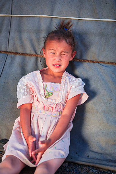 A young girl sits leaning against a canvas tent wall with a braided rope running horizontally behind her. She wears a pale pink dress with lace sleeves, ruffled trim and embroidered details; her hair is pulled into a small topknot with a blue clip. Squinting into bright sunlight, she holds her hands loosely clasped in her lap, with visible dust on her face and arms. Warm, directional light casts soft shadows on the fabric backdrop and the ground at her feet.