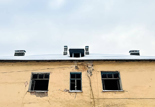 Snowy Rooftop Above Weathered Yellow Facade With Broken Windows, Climbing Dry Vines, Cracked Plaster, Peeling Paint And Small Chimney Cold Overcast Light Casts Muted Shadows Over Textured