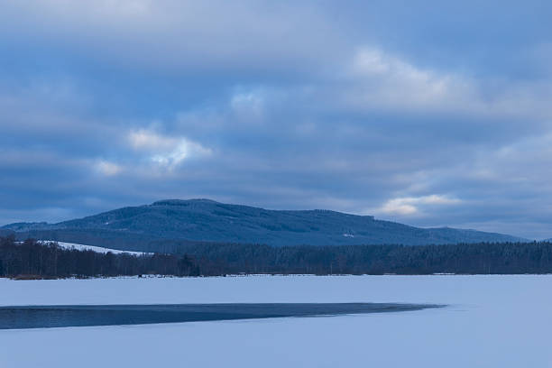 Winter landscape showing a partially frozen lake, snow covered forest, and mountains under a cloudy sky