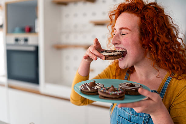 Joyful young red haired woman eating chocolate donut holding plate in bright modern kitchen
