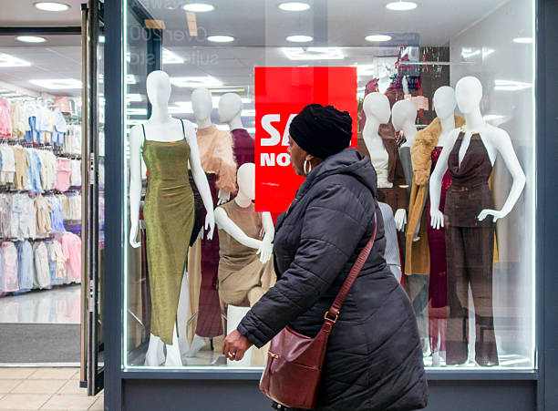 A woman walking past a shop window containing a collection of life-size mannequins displaying clothing, which are standing behind a red sale poster in a clothing store in the High Street, Bromley, Kent (Greater London).