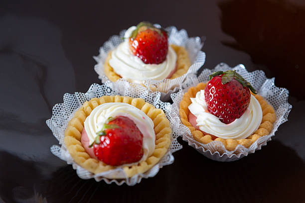 A group of individual strawberry tartlets presented against a dark, moody background to emphasize the vibrant red of the fruit.