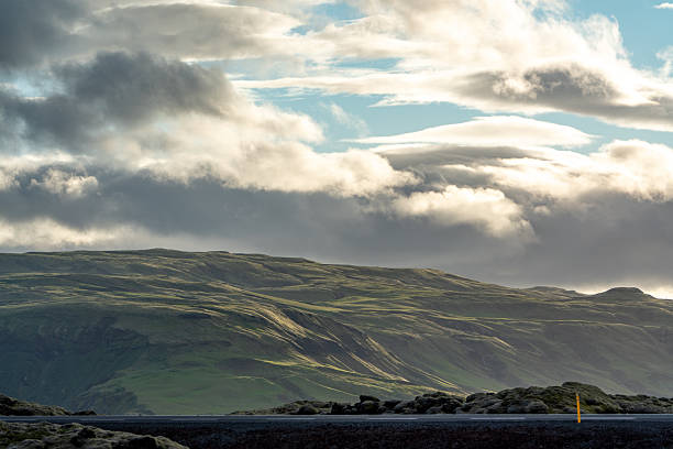 A vast, rolling green landscape under a dramatic sky filled with clouds. The sun breaks through the clouds, casting rays of light onto the hills, highlighting the textures and contours of the terrain. A road runs along the foreground, bordered by moss-covered rocks and dark volcanic soil, suggesting a remote and rugged location, possibly Iceland.