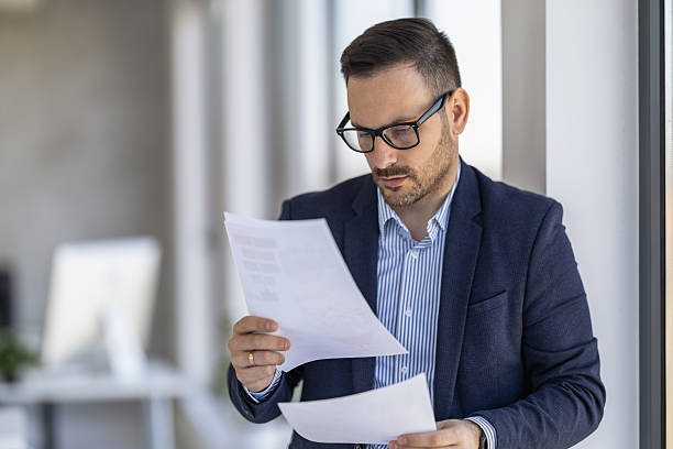 istock Professional Man Reading Documents By Window In Modern Office Attire And Glasses 2261296611
