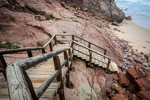 Weathered wooden stairs wind down from a red cliff toward a quiet beach