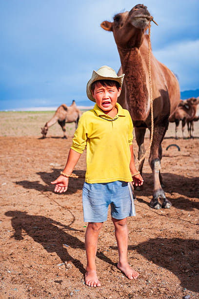 A barefoot young boy stands on a dry, sandy plain wearing a yellow shirt, light-blue shorts and a cowboy-style hat. He grips a rope tied around his wrist that is connected to a large camel standing close behind him; his face is scrunched as if crying or shouting. Other camels graze in the background beneath a wide blue sky, with long shadows stretching across the parched ground.