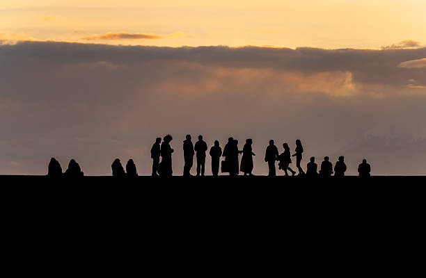 Silhouettes of a group of people walking on a concrete pier during a golden sunset. Dramatic mountain peaks and clouds in the background, Antalya Turkiye