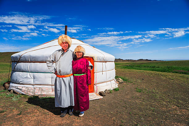 Two people stand arm-in-arm in front of a white felt ger (yurt) on an open grassy steppe. They wear traditional deels and furry hats — the person on the left in a light-grey robe with an orange sash and the person on the right in a deep magenta robe with a green sash. Behind them stretches a wide, gently rolling landscape under a vivid blue sky dotted with thin clouds, conveying a sense of rural nomadic life and calm openness.