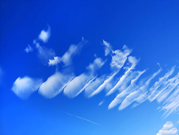 Fluffy clouds stretch across the blue sky, creating interesting shapes and patterns. The sunlight brightens the scene, and a jet trail is visible in the distance.
