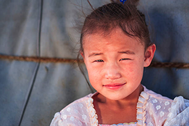 Close-up portrait of a young girl with her hair pulled back and a small blue hair tie. She squints against bright light, showing a faint, reserved smile and slightly flushed, freckled cheeks. She wears a pale, lace-trimmed dress with ruffled edges. The background is a muted fabric wall with a horizontal rope, and the image has warm, natural lighting and shallow depth of field that keeps focus on her face and expression.
