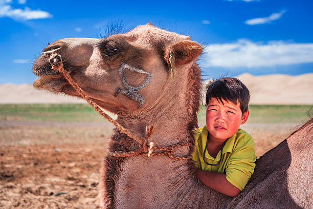 A close-up portrait of a young boy leaning against a camel. The camel's head fills the left side of the frame, showing a rope halter and a faded brand on its cheek. The boy, wearing a bright green shirt and a beaded necklace, looks toward the distance with sun-warmed, slightly flushed skin. Behind them are a flat, sandy plain and low sand dunes beneath a vivid blue sky dotted with a few clouds, creating a warm, open desert scene.