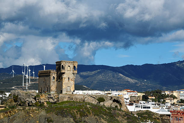 Tarifa, Spain: a landscape view featuring the Castillo de Santa Catalina in Tarifa, set against a backdrop of the town's white buildings and the mountain ranges of the Parque Natural del Estrecho. Visible in the distance is a modern wind farm, highlighting the region's status as a center for wind energy generation due to the strong Levante and Poniente winds. The image juxtaposes the early 20th-century historicist architecture of the castle with contemporary renewable energy