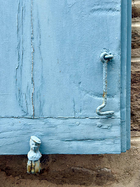 Close-up of weathered blue painted wooden shutter with metal latch and small decorative metal head detail at the bottom edge, set against a stone wall in Brittany, France.
