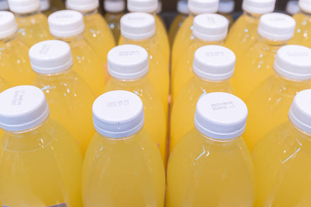 This high-angle close-up captures rows of clear plastic bottles filled with a bright yellow beverage, likely orange or lemon juice. The focus is sharp on the white screw-top caps, which are printed with black dot-matrix text indicating expiration dates (e.g., \