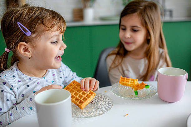 Two young girls sit at the table having breakfast, representing warmth, routine, and family bonding.