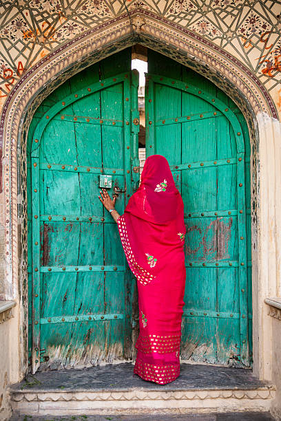 A woman seen from behind wears a vibrant magenta-red embroidered sari and rests a henna-adorned hand on a large, weathered turquoise wooden door. The arched doorway is framed by faded floral and geometric frescoes and studded metal fittings; peeling paint, scuffed steps and a locked latch add a textured, timeless quality. The strong color contrast between the sari and the door creates a striking, photographic composition.