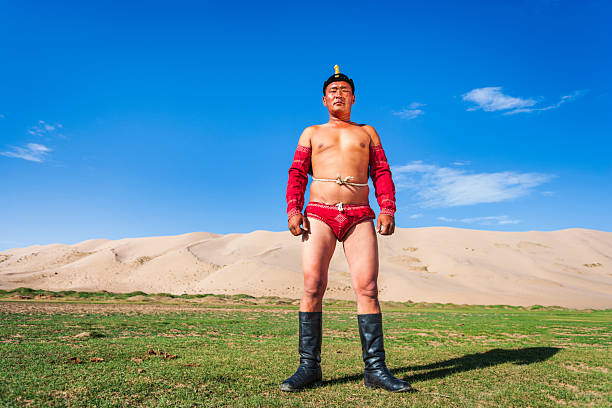 A shirtless man stands confidently in the center of the frame wearing bright red traditional wrestling garments (arm sleeves and briefs), a small decorated hat, a rope belt, and tall black boots. He poses on a patch of green grass with sweeping pale sand dunes behind him under a vivid blue sky dotted with a few clouds. Strong sunlight casts a clear shadow and highlights the textures of his clothing, skin and the desert landscape, creating a bold, high-contrast portrait with cultural and athletic overtones.