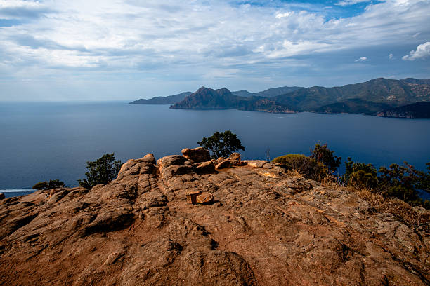 Rocky plateau overlooking the sea and rugged coastline with distant mountains under a partly cloudy sky in Corsica, France. Ideal for travel, coastal landscape, and nature backgrounds.