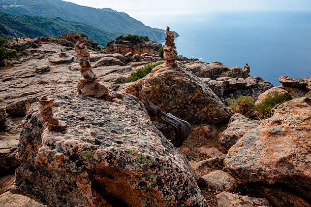 Stone cairns arranged on a rocky coastal plateau with cliffs, sea, and distant hills under a bright cloudy sky, suitable for travel, hiking, and outdoor destination concepts.