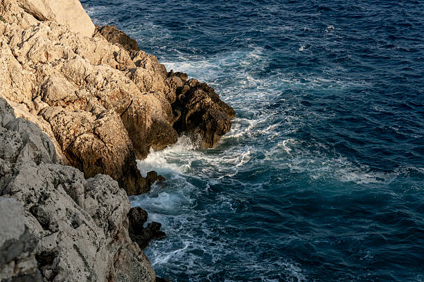 Powerful waves crash against rugged coastal rocks under warm natural light. Deep blue sea water swirls around textured stone formations, creating a dynamic Mediterranean seascape with strong contrast between golden cliffs and turquoise water. No people.