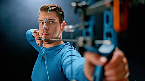 Caucasian archer in an indoor setting focuses on perfecting his bow technique with precision and concentration. Holding a modern bow, preparing to release an arrow, aiming for accuracy and success in the target shot.