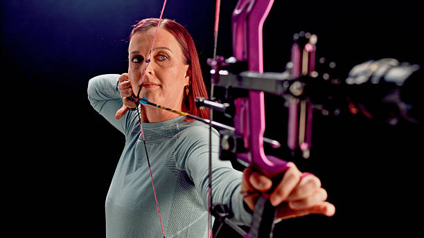 Mature Caucasian female archer in an indoor range focusing intently on her target with a purple compound bow. Demonstrating concentration and precision in her archery technique.