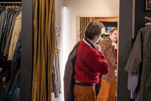 Male customer in men's wear second hand boutique trying on vintage fashionable checkered blazer in front of mirror in open fitting room. Mustachioed elegant man in jumper fits on jacket in shop.