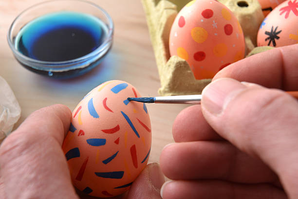 Close-up of adult hands painting a pastel Easter egg with colorful patterns. Creative DIY activity with blue dye and egg carton in background.