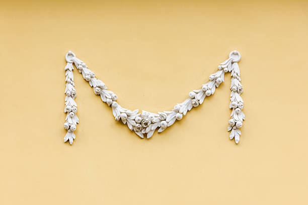 Elegant white plaster garland mounted on a soft yellow wall, featuring ornate floral molding and classic architectural detail. Minimal composition emphasizes texture, symmetry and vintage charm