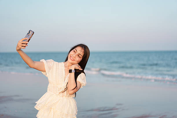Beautiful asian woman in white dress enjoying while taking selfie with smartphone on the seaside beach. creating joyful travel and leisure atmosphere