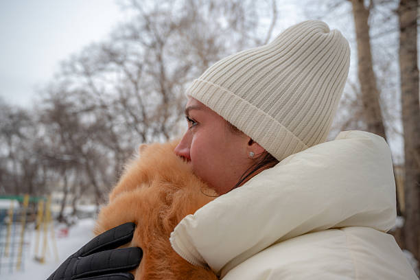 A young girl in a cream-colored hat and a voluminous down jacket holds a red dog tenderly in her arms and embraces it. They are on a winter street: in the background there are bare trees and a playground, the sky is overcast with clouds. The girl's gaze is thoughtful, the atmosphere of the picture is calm, filled with comfort and warmth of friendly affection despite the coolness around.