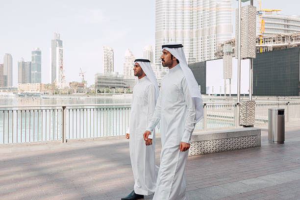 Two Middle Eastern men walk side by side along a waterfront promenade while talking. The relaxed outdoor setting reflects an informal business discussion, networking, and idea sharing in a contemporary urban environment.