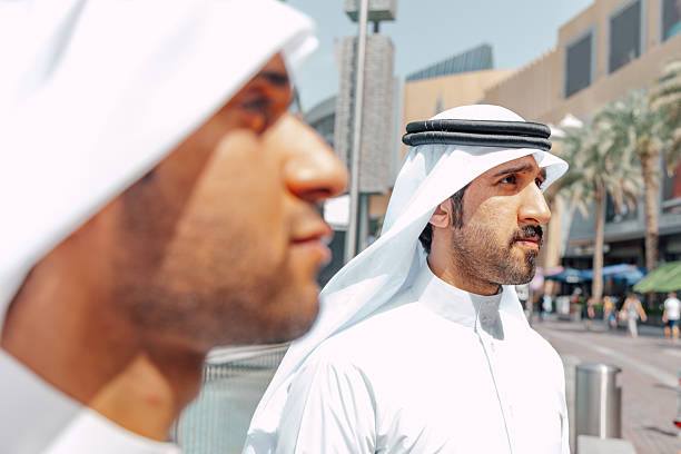 Two Arab businessmen wearing traditional white clothing greet each other outdoors in a modern city setting. The handshake symbolizes trust, agreement, and the start of a professional partnership, combining cultural heritage with contemporary business values.