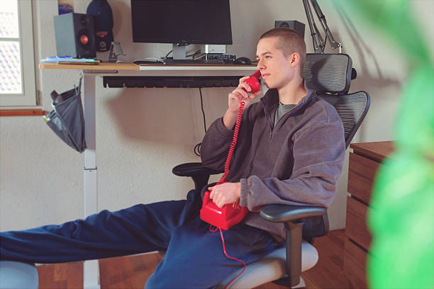 Teen boy leaning back comfortably in an office chair, speaking on a bright red vintage landline phone. Warm interior tones and negative space around the subject provide room for copy.