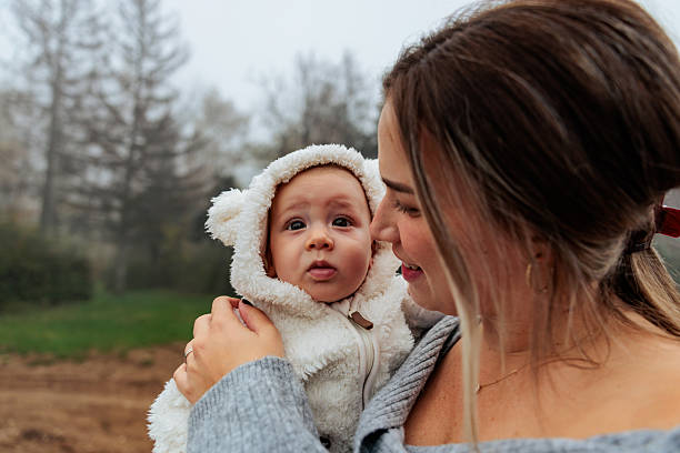 Close portrait of a young mother holding her baby in her arms outdoors on a misty day. The infant wears a soft hooded outfit with bear ears and looks toward the camera, while the mother smiles warmly at the child.