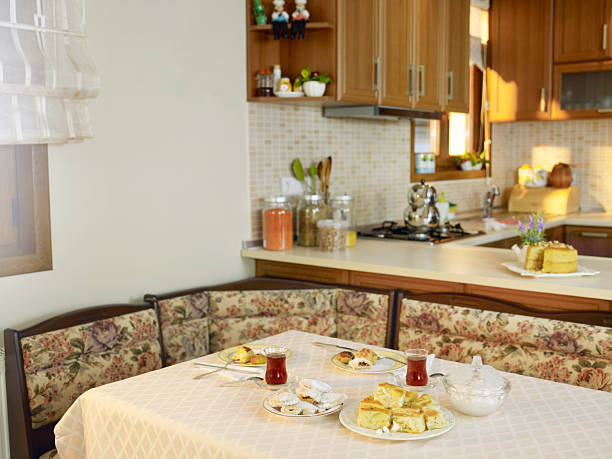 A cozy domestic kitchen features a table set with tea, pastries, and a lace tablecloth in front of a cushioned bench, with wooden cabinets and warm lighting in the background. This inviting home interior scene is ideal for family lifestyle themes, hospitality concepts, food and drink articles, and home decor promotions. Horizontal composition with copy space suitable for magazines, web banners, and social media storytelling.