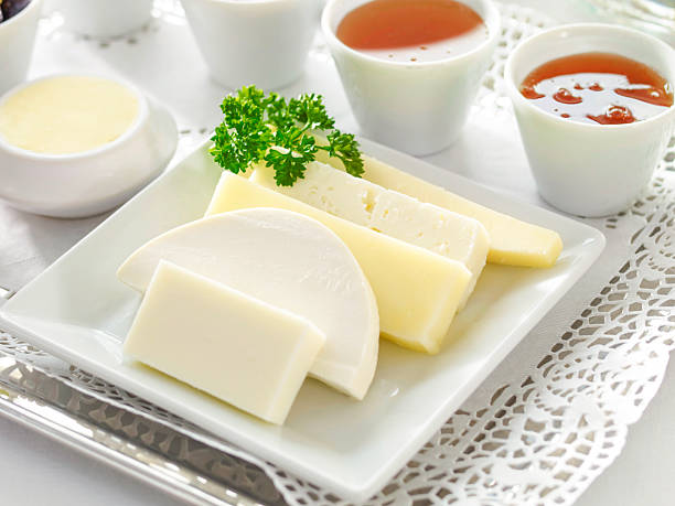 Close up view of a breakfast plate featuring assorted white cheeses neatly arranged on a square white tray, garnished with fresh parsley and surrounded by small bowls of sauces and spreads on a lace placemat. The bright morning food scene on a white background with copy space is ideal for hotel breakfast promotions, healthy dairy concepts, Mediterranean or Turkish breakfast themes, and cafe menu designs. Horizontal high resolution composition works well for cookbooks, food blogs, catering brochures, and social media posts showcasing elegant breakfast presentations.​