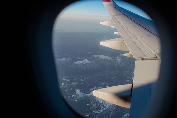 View from an aircraft window showing an airplane wing flying above the snow-covered peaks of the Alps under a clear blue sky. Travel, transportation, and adventure concept.