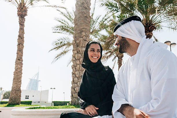 An Emirati man and woman sit together on a bench in a park surrounded by palm trees, smiling and talking, representing calm moments, tradition, and modern family life.