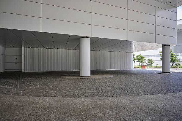 A building structure features white walls and pillars. The area is open and has a paved ground. Trees are visible in the background under clear sunshine.