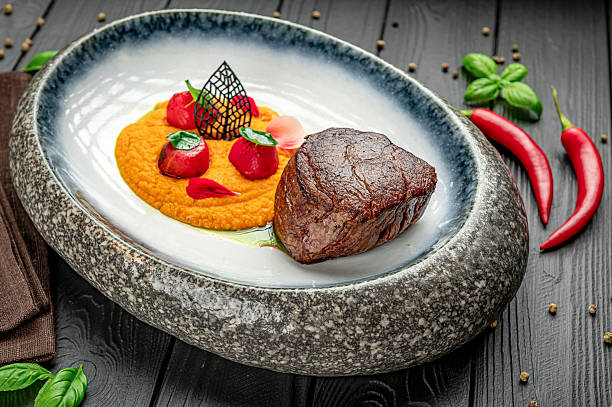 Fine dining dish with meat and vegetables served on unique plate design. A gourmet dish features cooked meat with vegetable puree and sauces arranged artistically on a stone plate.